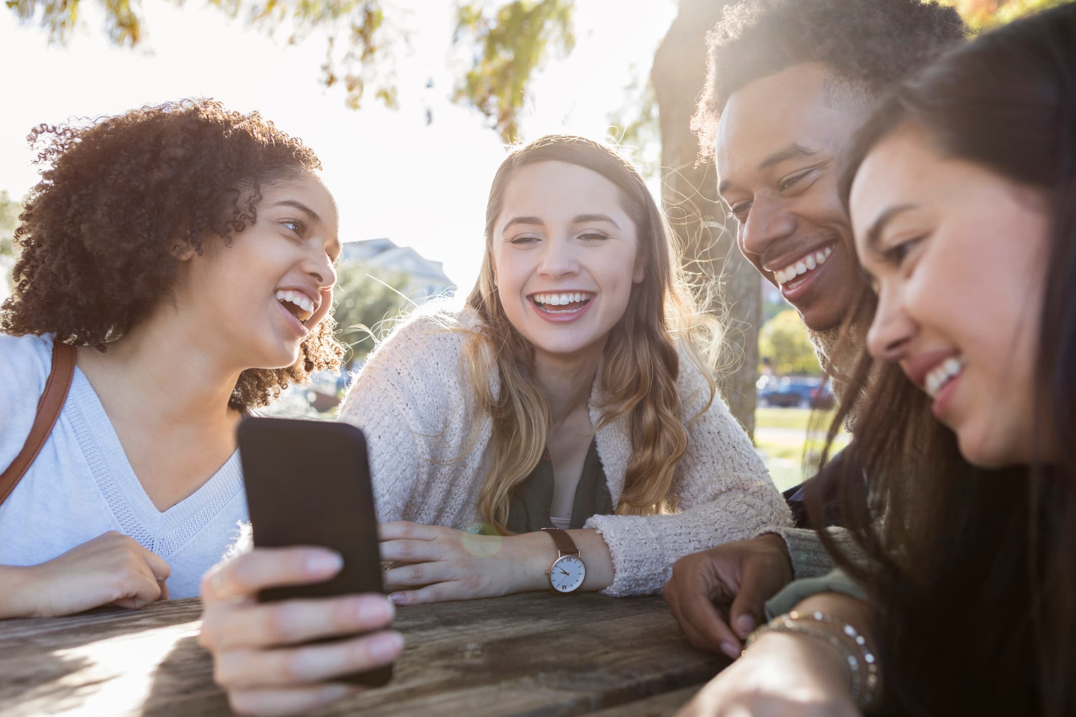 A woman sat outside at a table holds a smartphone in her right hand. A man to her left and a woman to her left look at the screen of her phone, while a woman to her right looks at the man.