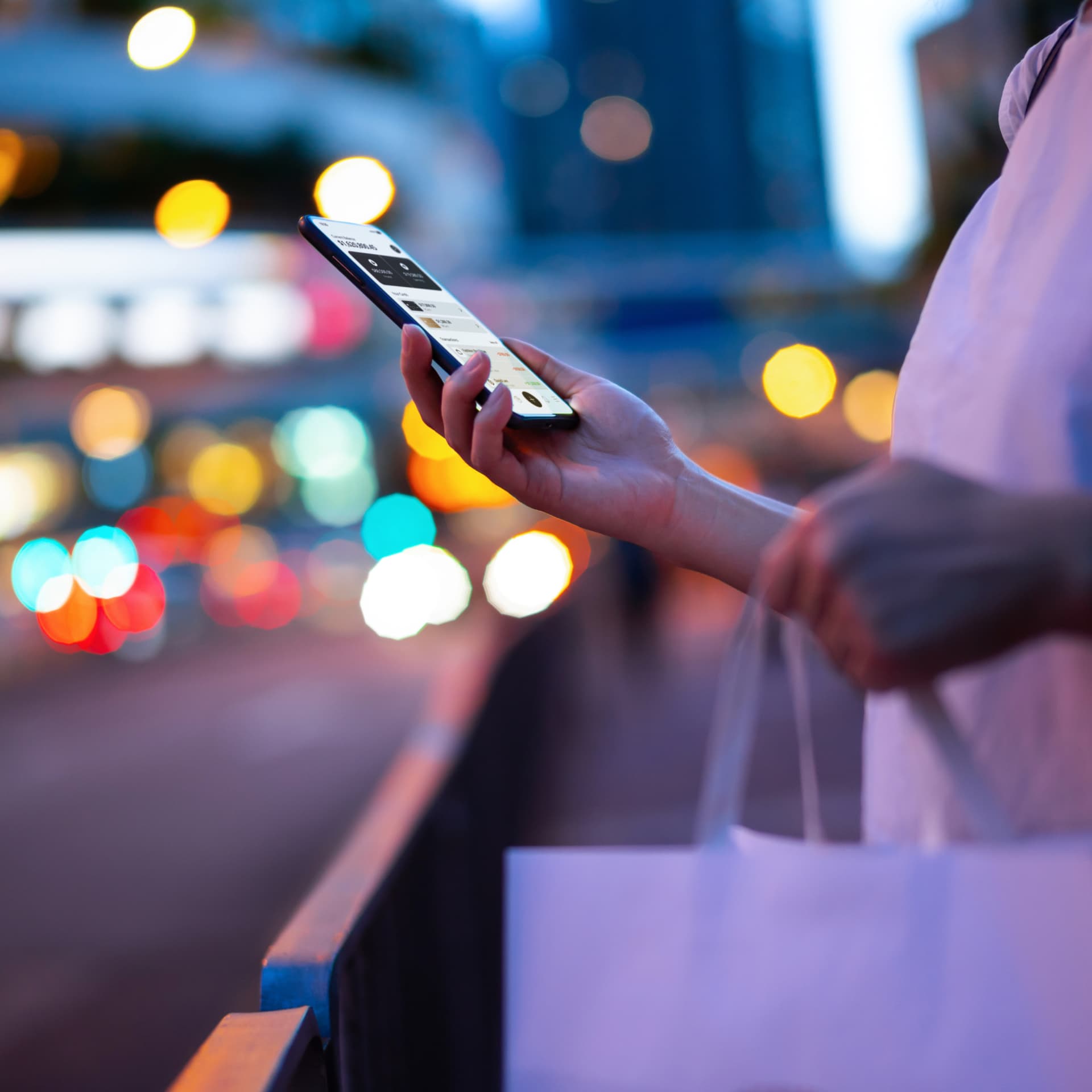 Close up photo of woman carrying a paper shopping bag, looking at her smartphone in city street scape with illuminated lights