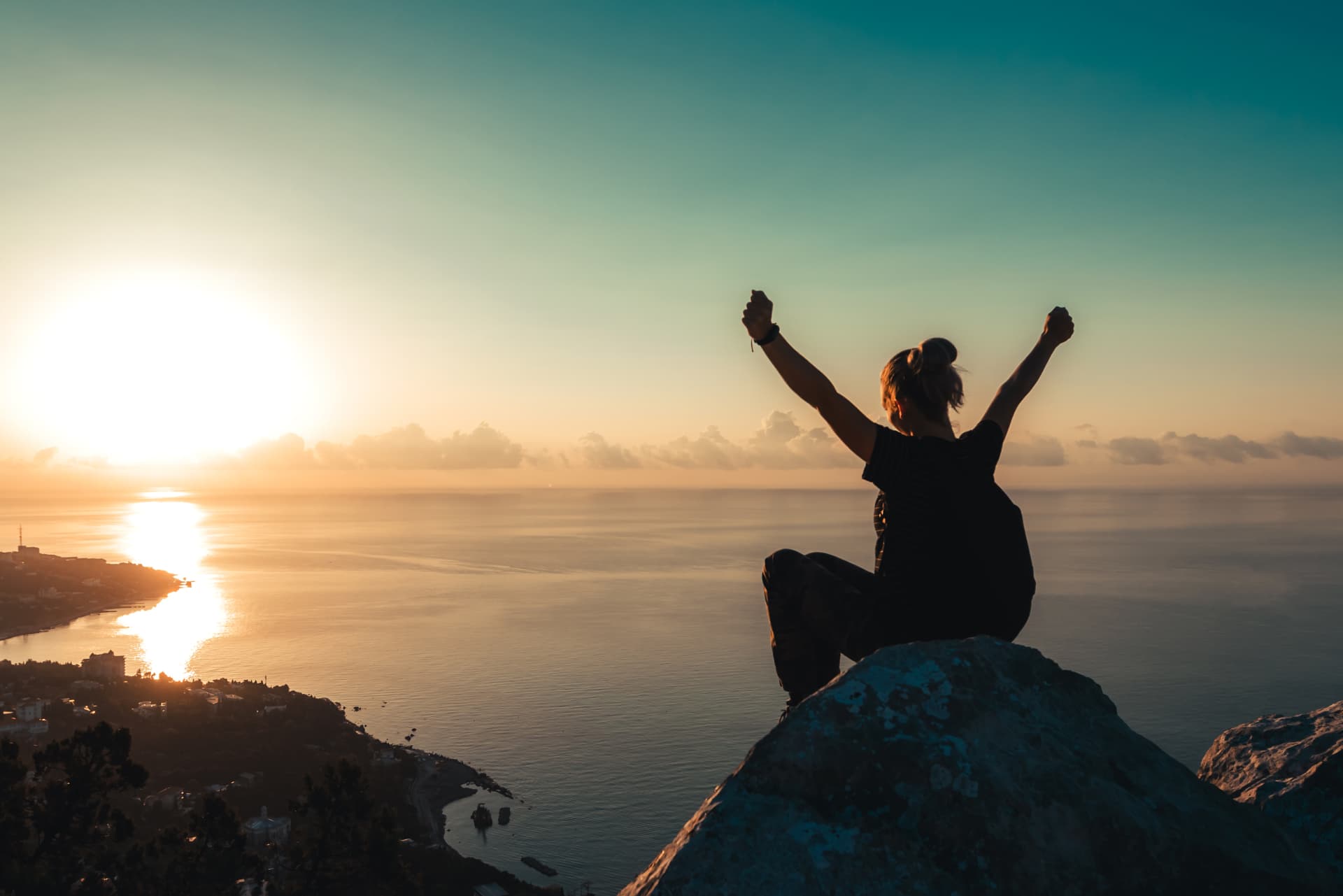 Girl sitting on top of mountain with arms up in celebration watching sunset