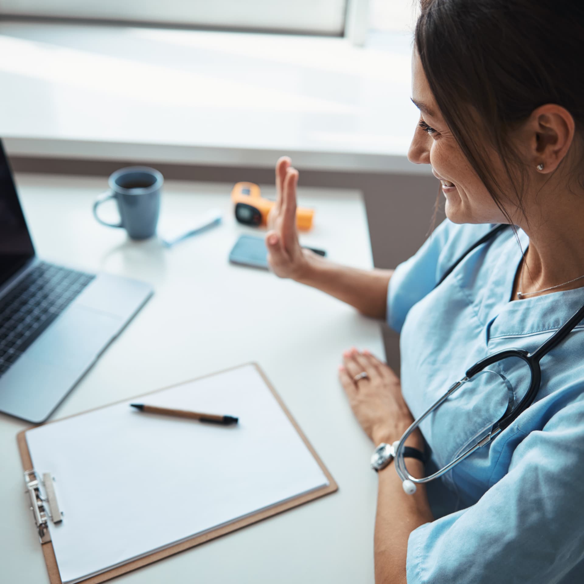 Joyful woman physician doing hello gesture and smiling while sitting at the table with notebook and giving online consultation